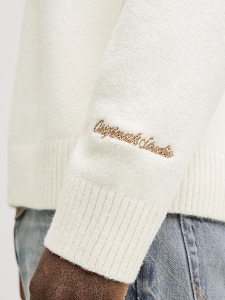 Close-up of a cream-colored sweater with gold 'Originals Studio' embroidery on a white background