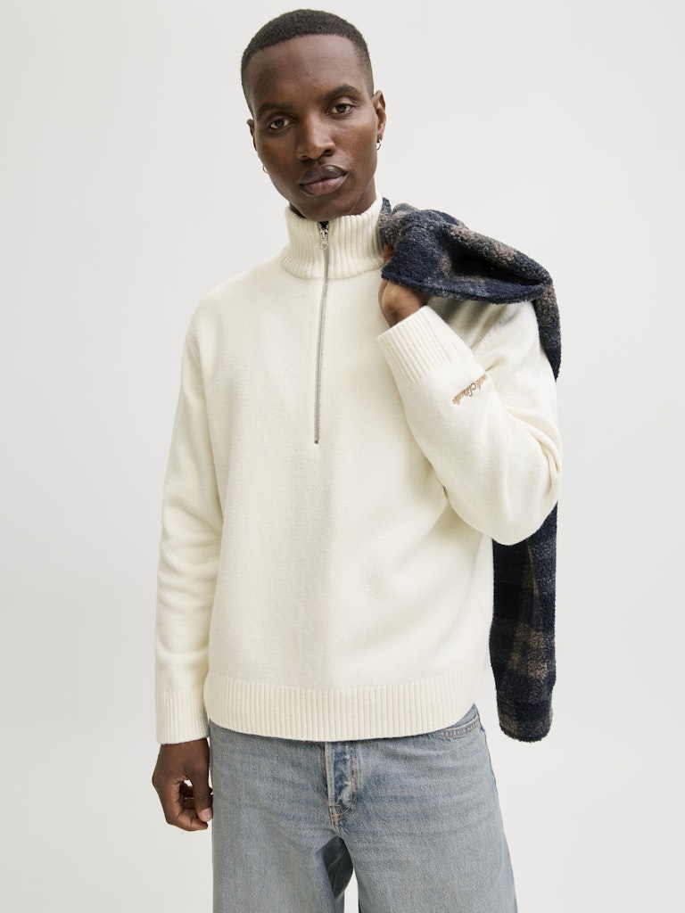 Man wearing a cream half-zip sweater holding a dark jacket against a light gray background