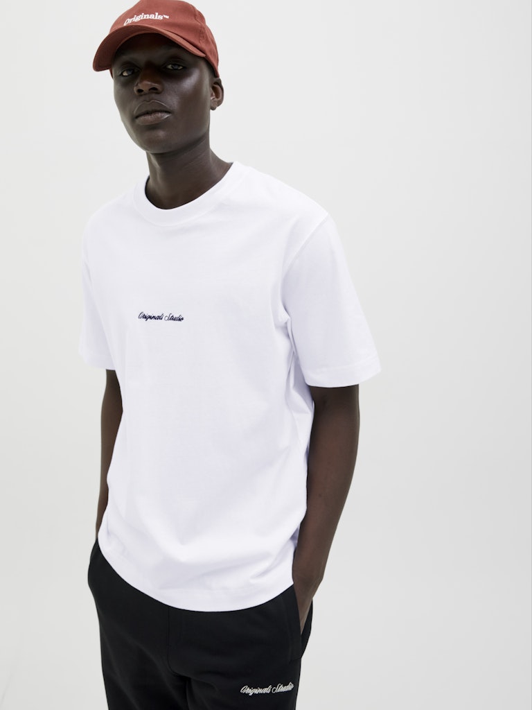 Person wearing a white t-shirt with a logo and black pants, standing against a plain background