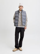 Person wearing a gray puffer vest over a beige shirt, black pants, and a white beanie on a white background