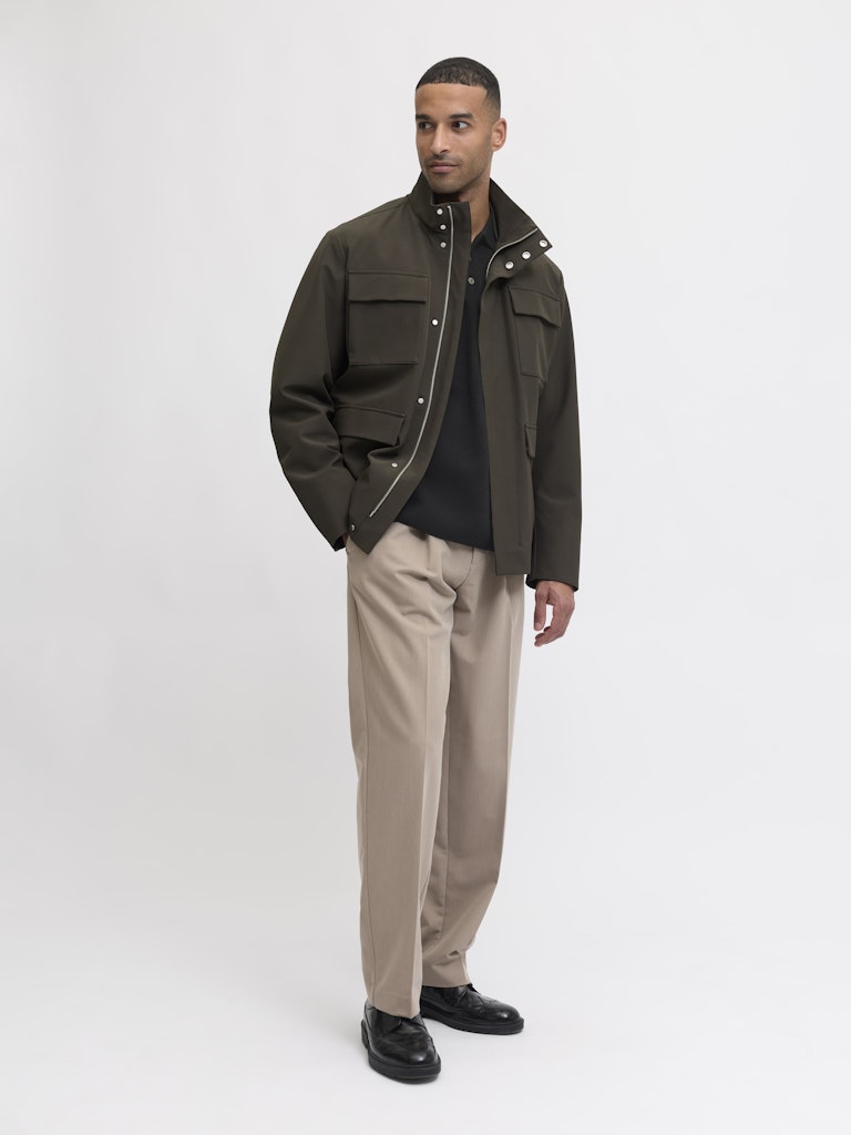Man wearing a green jacket, black shirt, beige pants, and black shoes on a white background