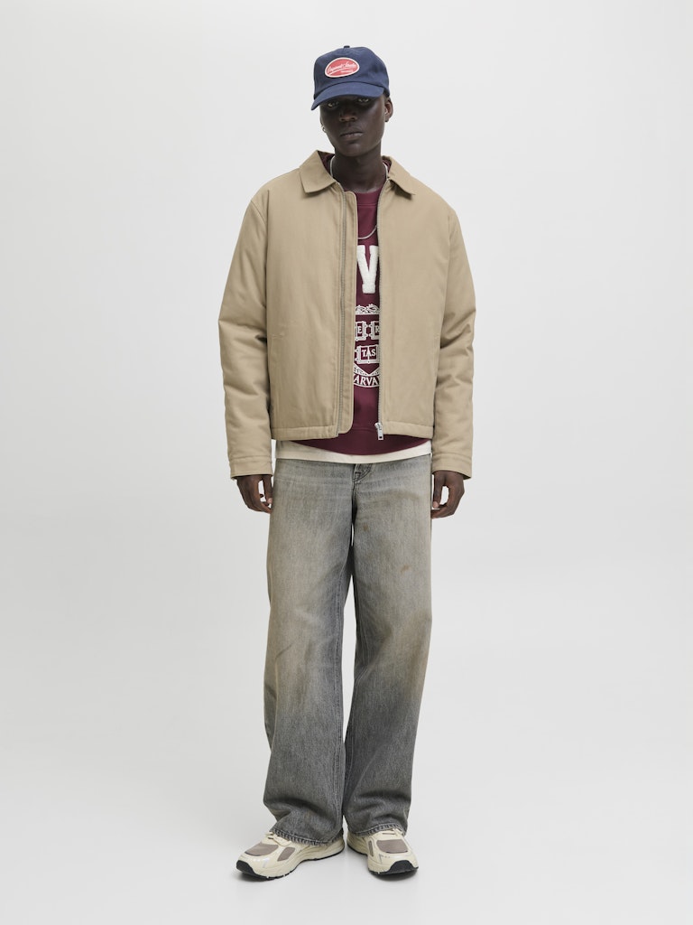 Person wearing a beige jacket, maroon shirt, and blue jeans on a white background