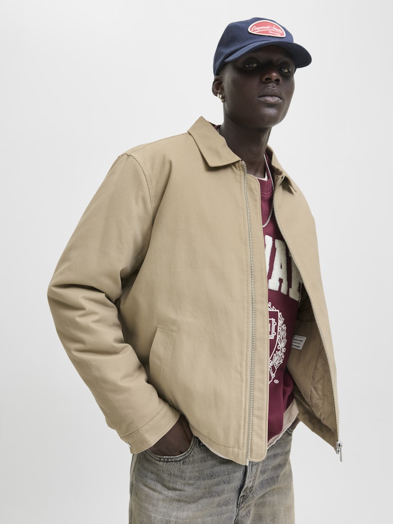 Person wearing a beige jacket and maroon shirt with a cap on a white background