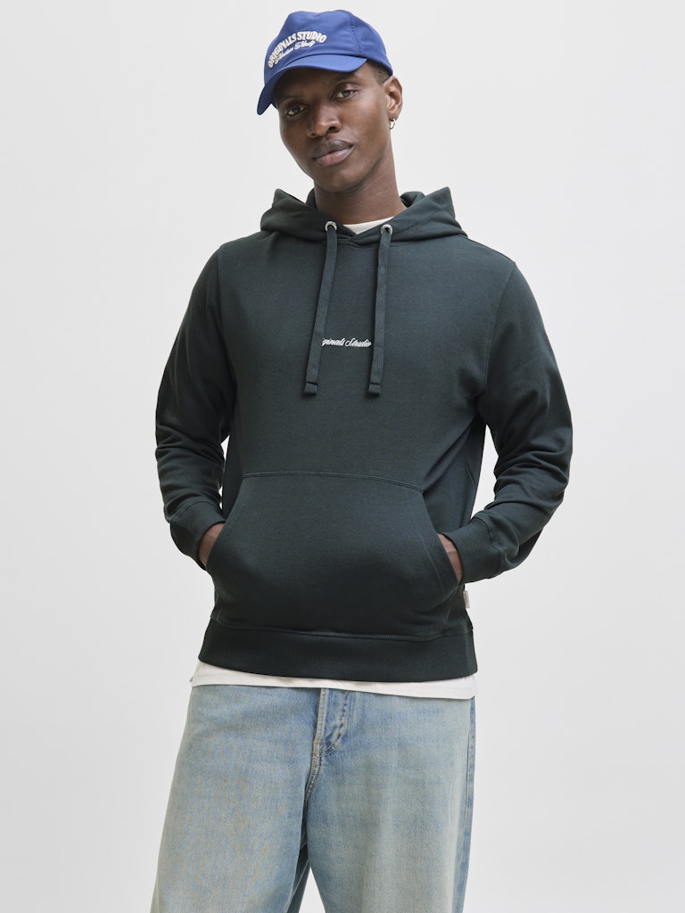 Person wearing a dark green hoodie, blue jeans, and a blue cap on a light gray background