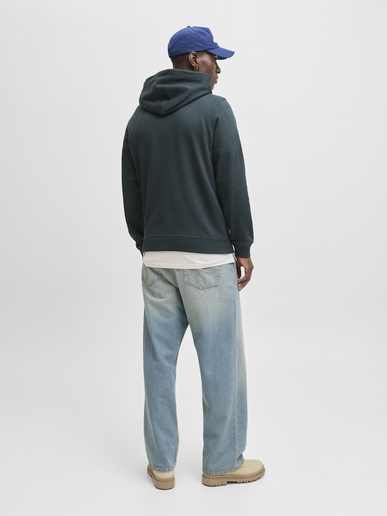 Person wearing a dark green hoodie, light blue jeans, and a blue cap on a white background
