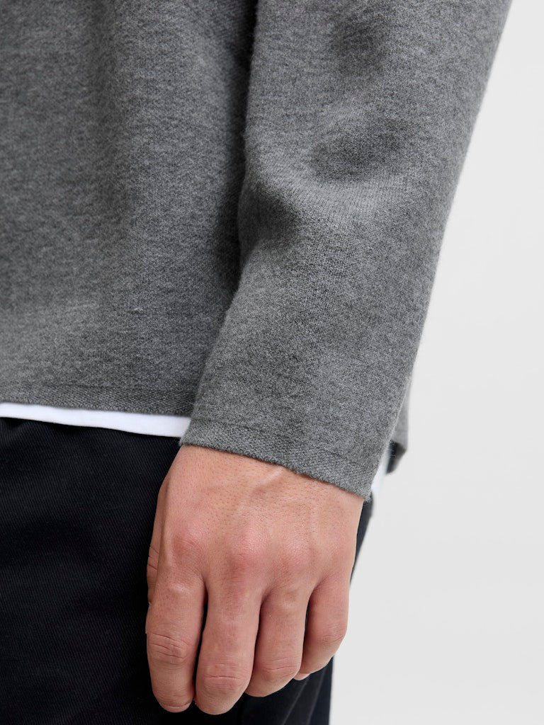 Close-up of a person wearing a gray sweater with a white shirt underneath on a light gray background.