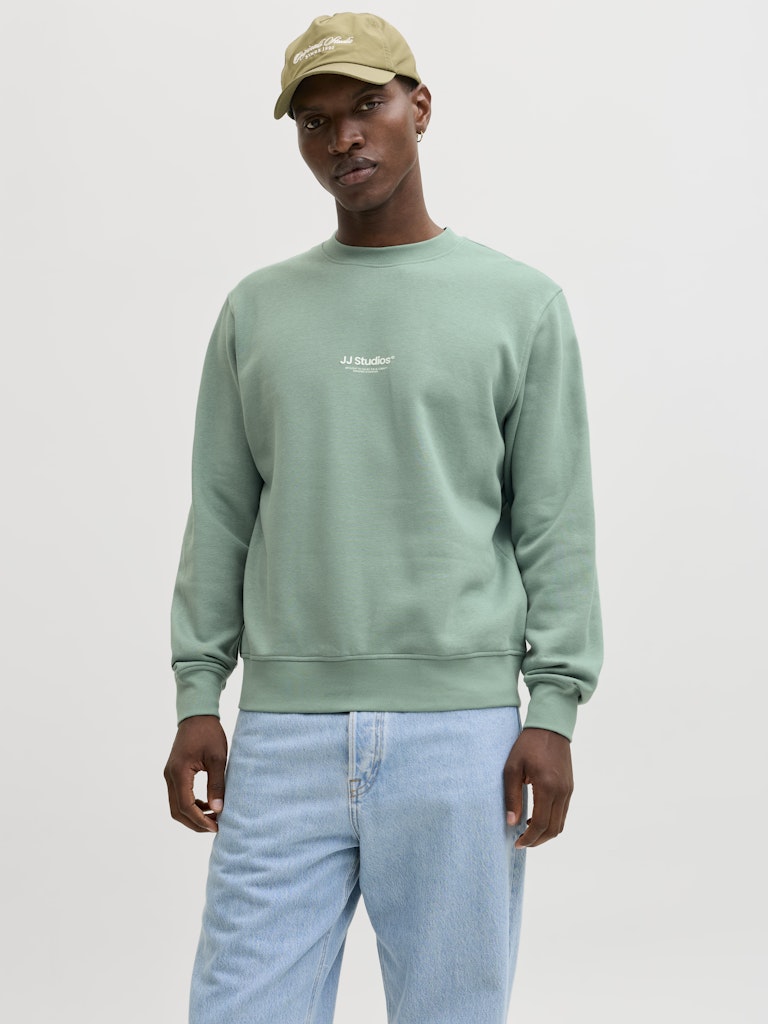 Man wearing a green sweatshirt and light blue jeans on a plain background