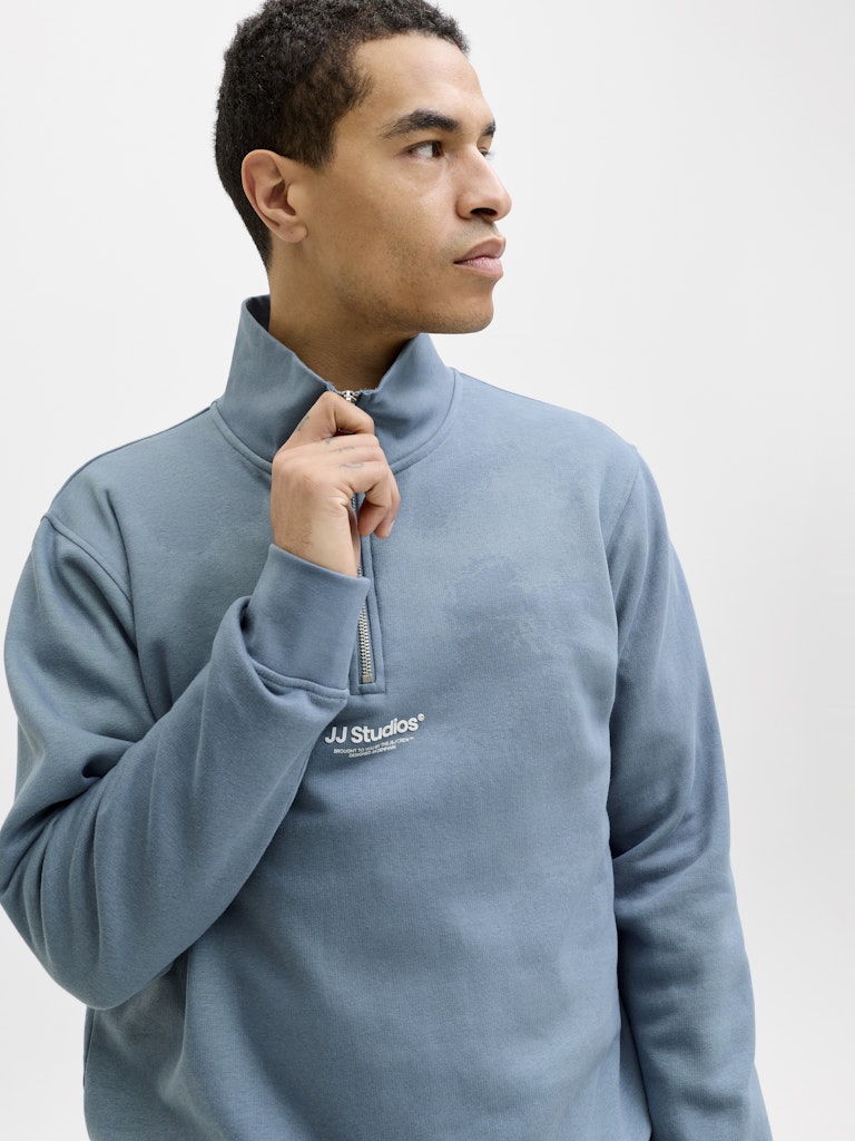 Man wearing a blue sweatshirt with quarter zip and 'JJ Studios' logo on a white background