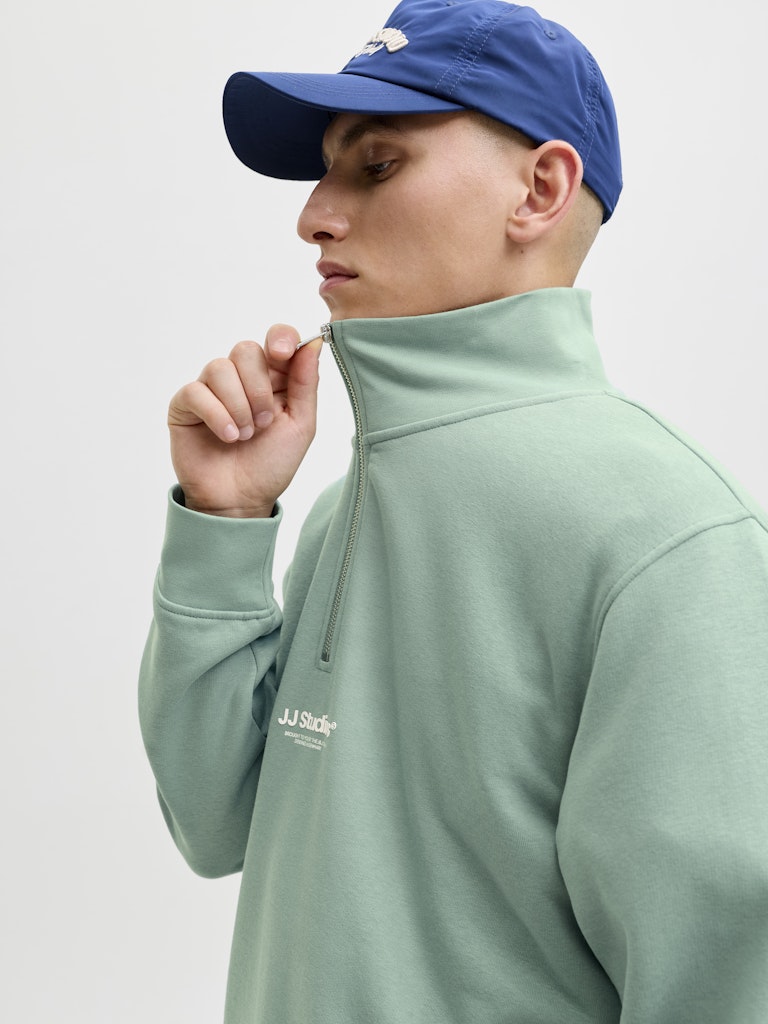 Man wearing a green half-zip sweatshirt and blue cap on a light gray background