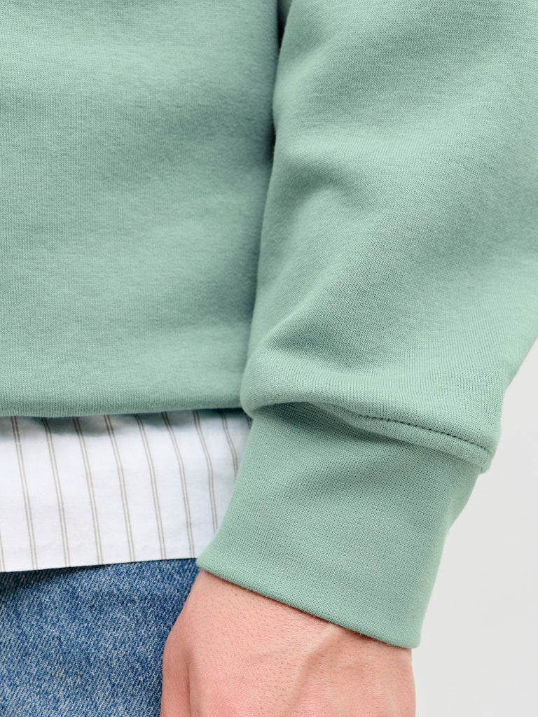 Close-up of a person wearing a light green sweatshirt with a white shirt and blue jeans underneath.