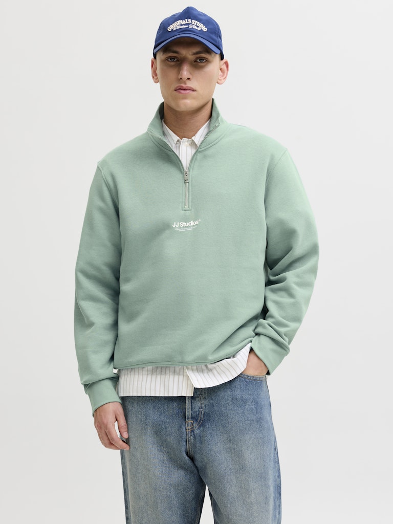 Person wearing a green sweater, blue jeans, and a blue cap on a white background