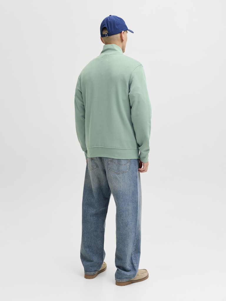 Person wearing a green sweater and blue jeans standing against a white background