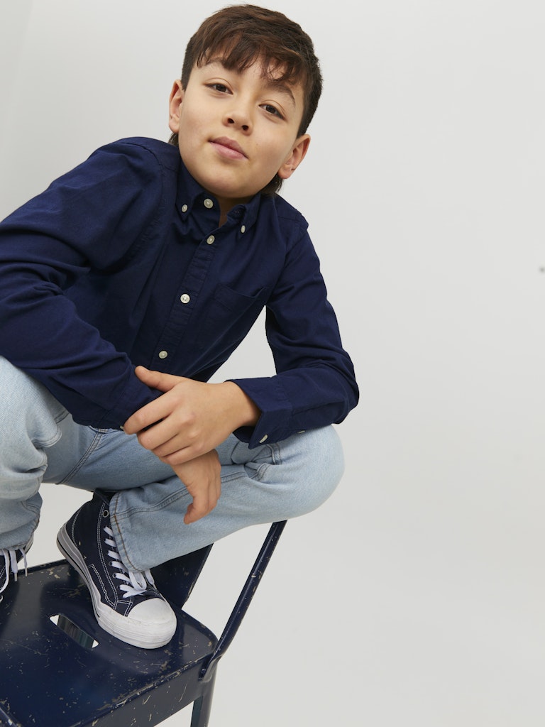 Young boy wearing a navy shirt and light blue jeans sitting on a black stool against a white background