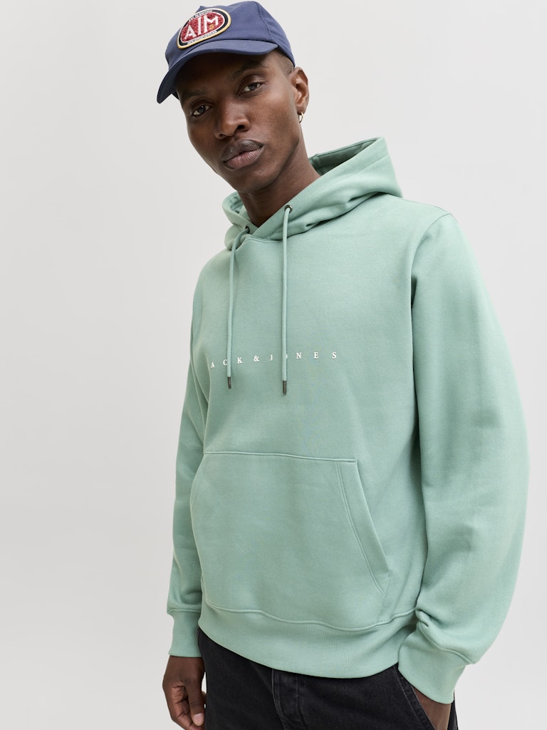 Person wearing a light green hoodie with text and a navy cap with a logo on a white background
