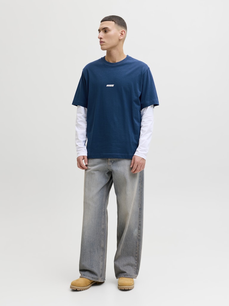 Man wearing a blue t-shirt with a white logo, white long-sleeve shirt, gray jeans, and tan shoes on a white background