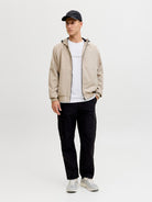 Man wearing a beige jacket, white shirt, black pants, and cap on a light gray background