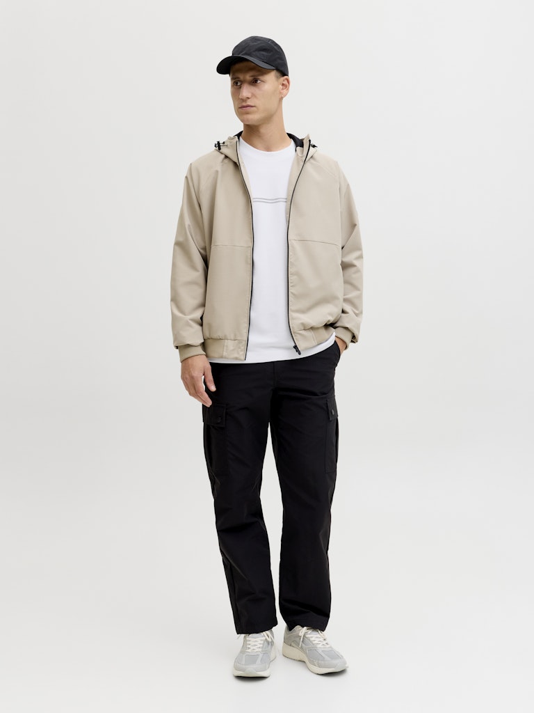 Man wearing a beige jacket, white shirt, black pants, and cap on a light gray background