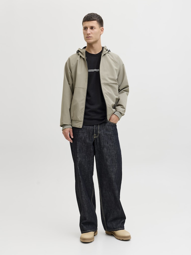 Man wearing a beige jacket, black t-shirt, and dark jeans on a white background