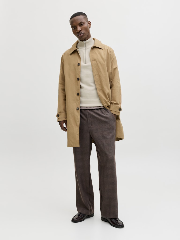 Man wearing a beige trench coat over a white sweater and brown pants on a white background