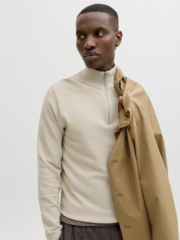 Man wearing a cream knit half zip sweater with a jacket over one shoulder