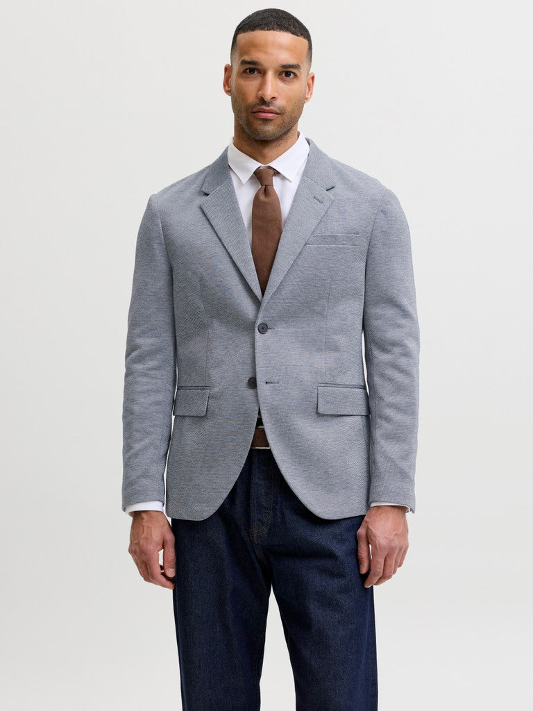 A man wearing a light gray textured blazer over a white dress shirt and brown tie, paired with dark blue jeans, standing against a light gray studio background.