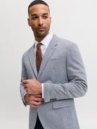 A man wearing a light gray heathered suit jacket, a white dress shirt, and a brown tie, posing against a light gray studio background.