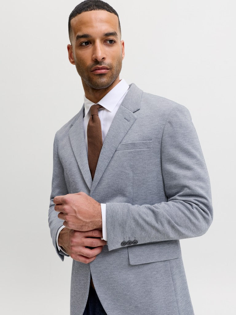 A man wearing a light gray heathered suit jacket, a white dress shirt, and a brown tie, posing against a light gray studio background.