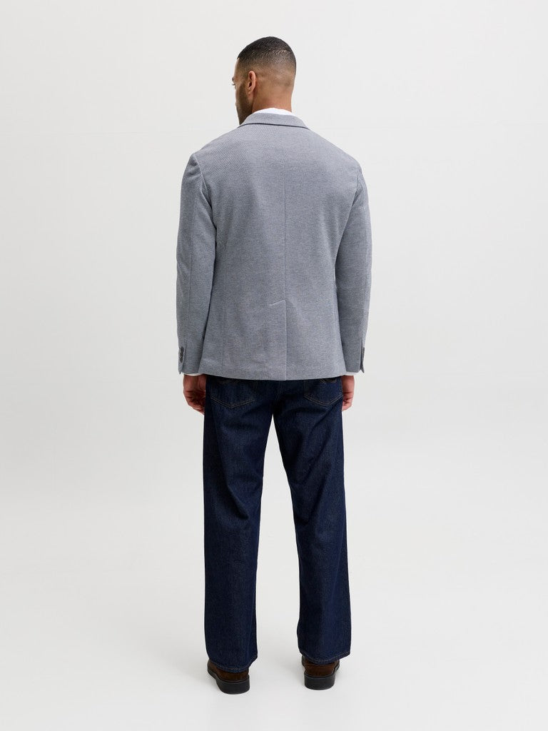 A man viewed from the back wearing a light gray blazer and dark navy trousers, standing against a plain light gray studio background.