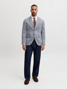 A man modeling a light gray textured blazer, dark navy trousers, a white dress shirt, a brown tie, and brown loafers, standing against a plain light gray studio background.