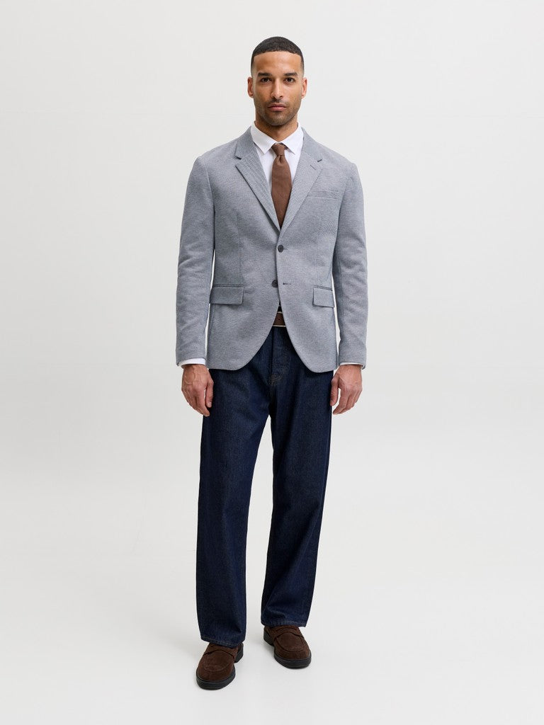 A man modeling a light gray textured blazer, dark navy trousers, a white dress shirt, a brown tie, and brown loafers, standing against a plain light gray studio background.