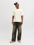 Man wearing a yellow t-shirt, dark jeans, and a cap on a light gray background