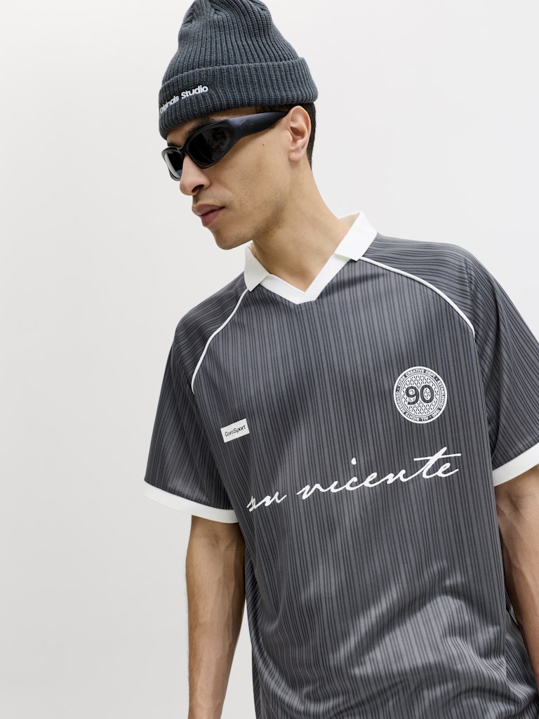 Person wearing a gray sports jersey with white accents and text, sunglasses, and a beanie on a light gray background.