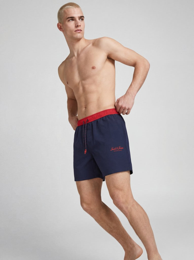 Man wearing navy swim shorts with a red waistband on a gray background