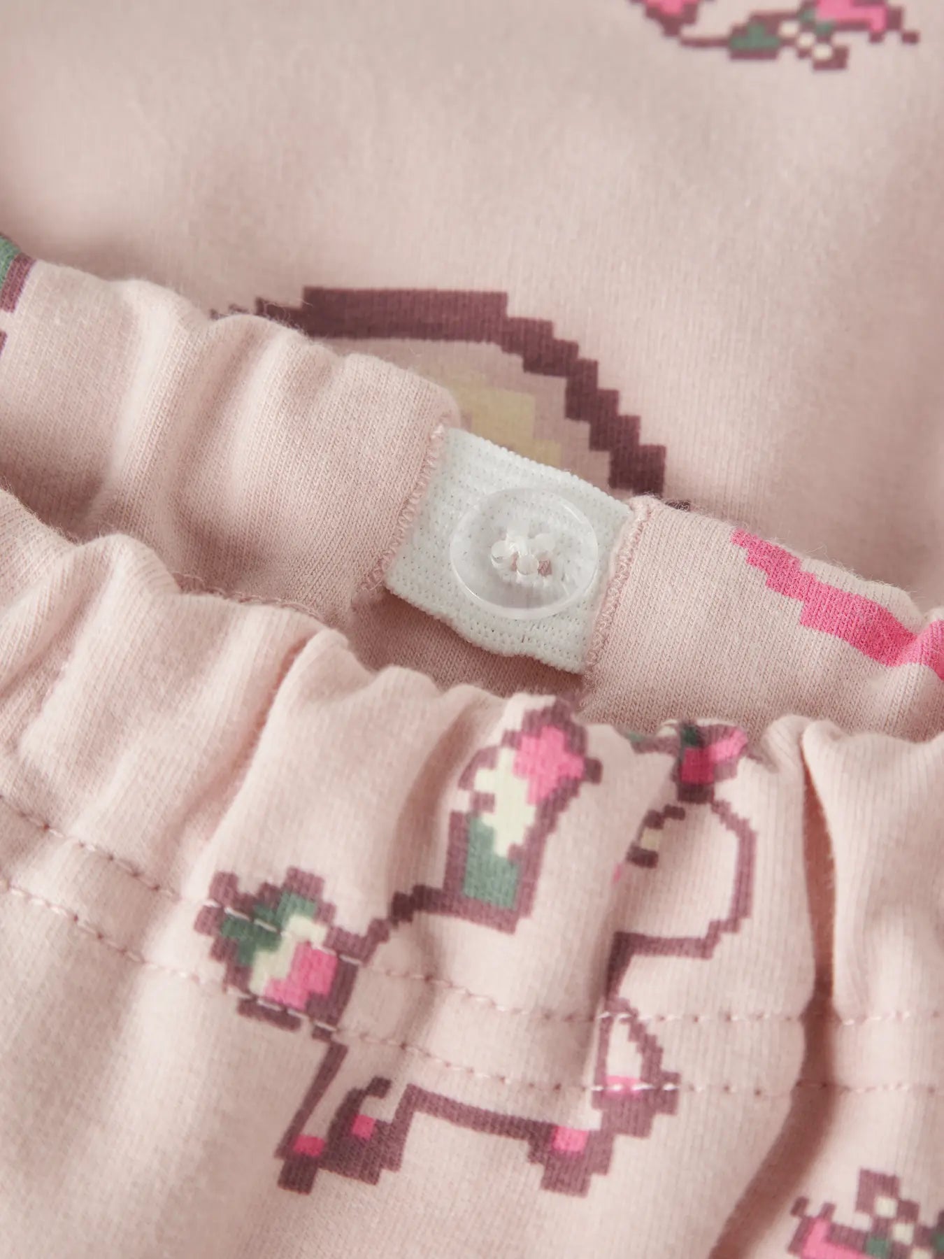 Close-up of a light pink fabric garment with a pixelated unicorn pattern in pink, purple, and green, featuring a white elastic waistband with a snap button.