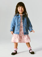 A young girl smiling and posing in a studio setting, wearing a light blue denim jacket over a pink tiered dress with a unicorn print, paired with pink socks and black shoes, against a light gray background.