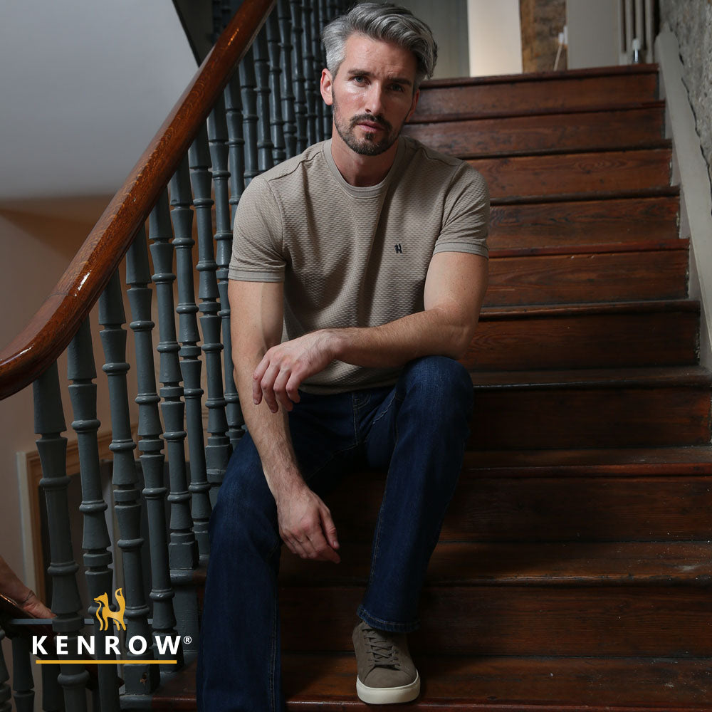 Man sitting on a wooden staircase wearing a brown t-shirt and blue jeans, with 'KENROW' logo visible.