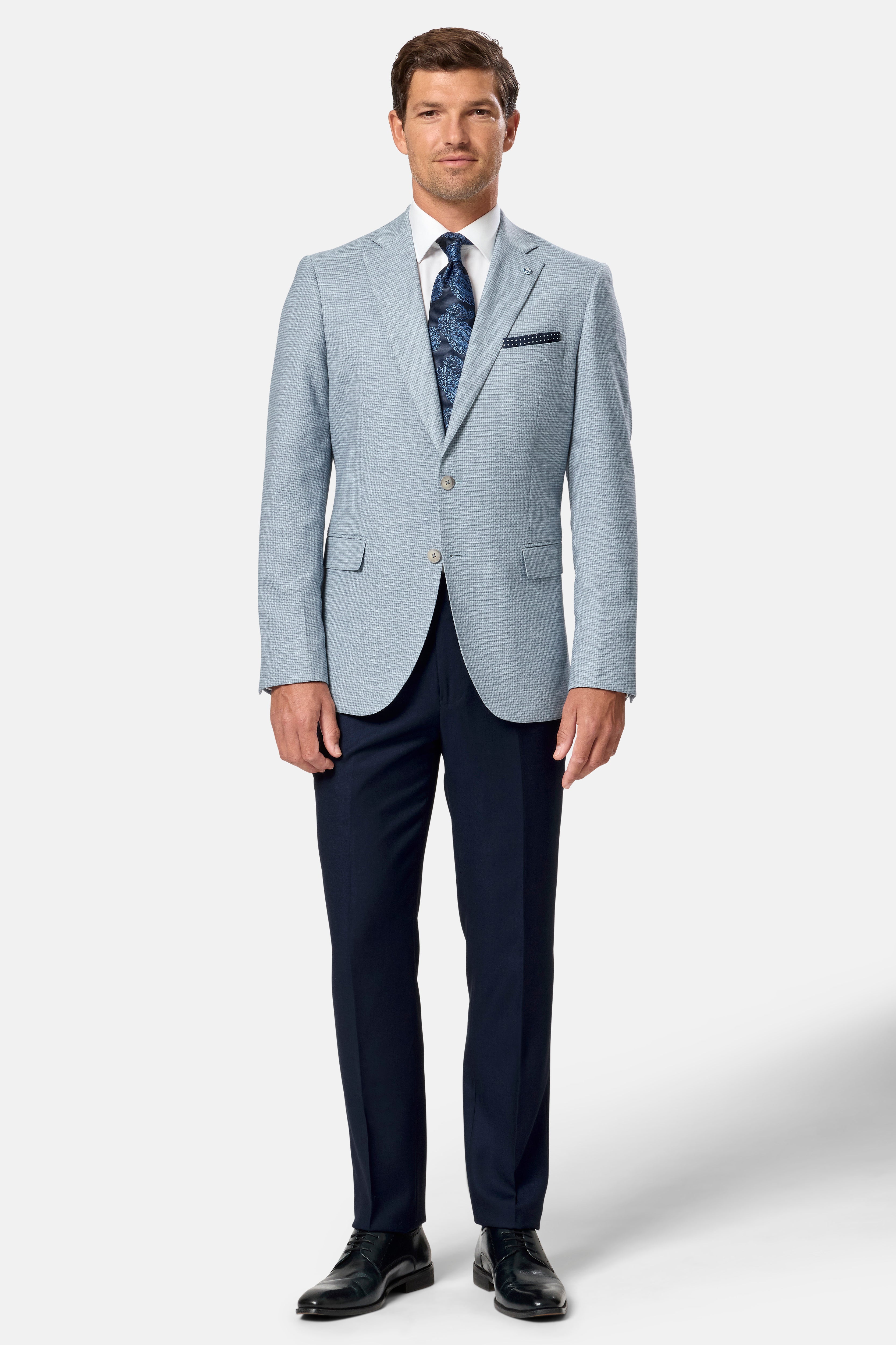 Man wearing a light blue blazer over a navy dress shirt with dark blue pants on a white background