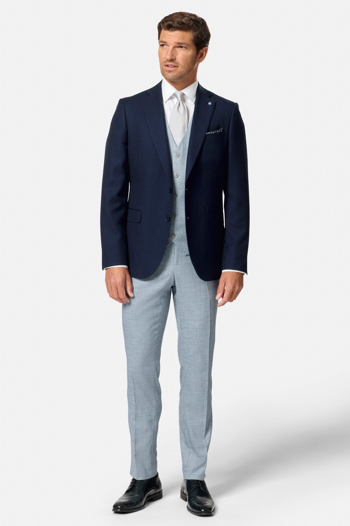 Man wearing a navy blazer, light blue vest, and pants on a white background