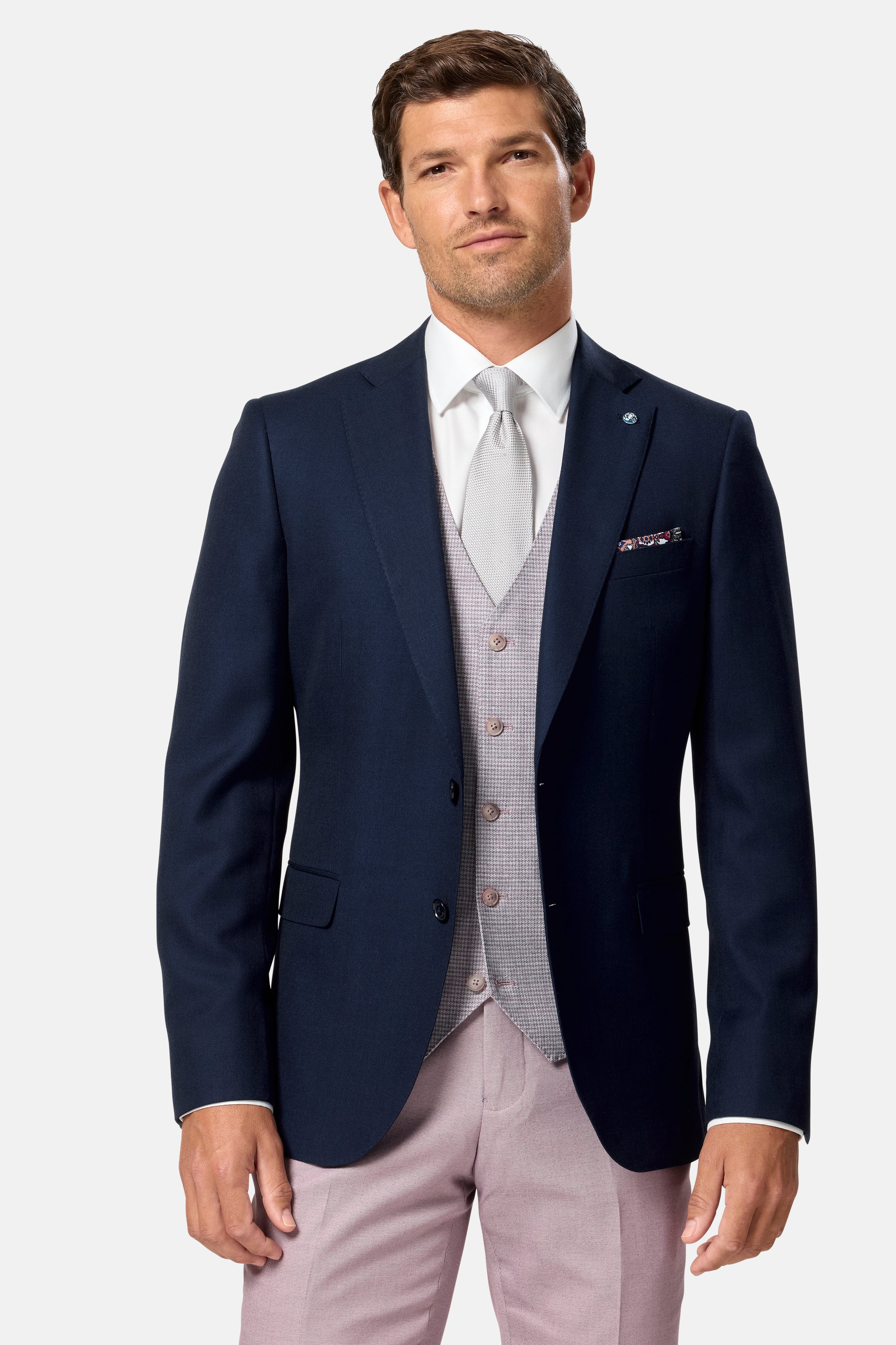 Man wearing a navy blazer blush check waistcoat, white shirt, gray vest, and pink pants on a light gray background