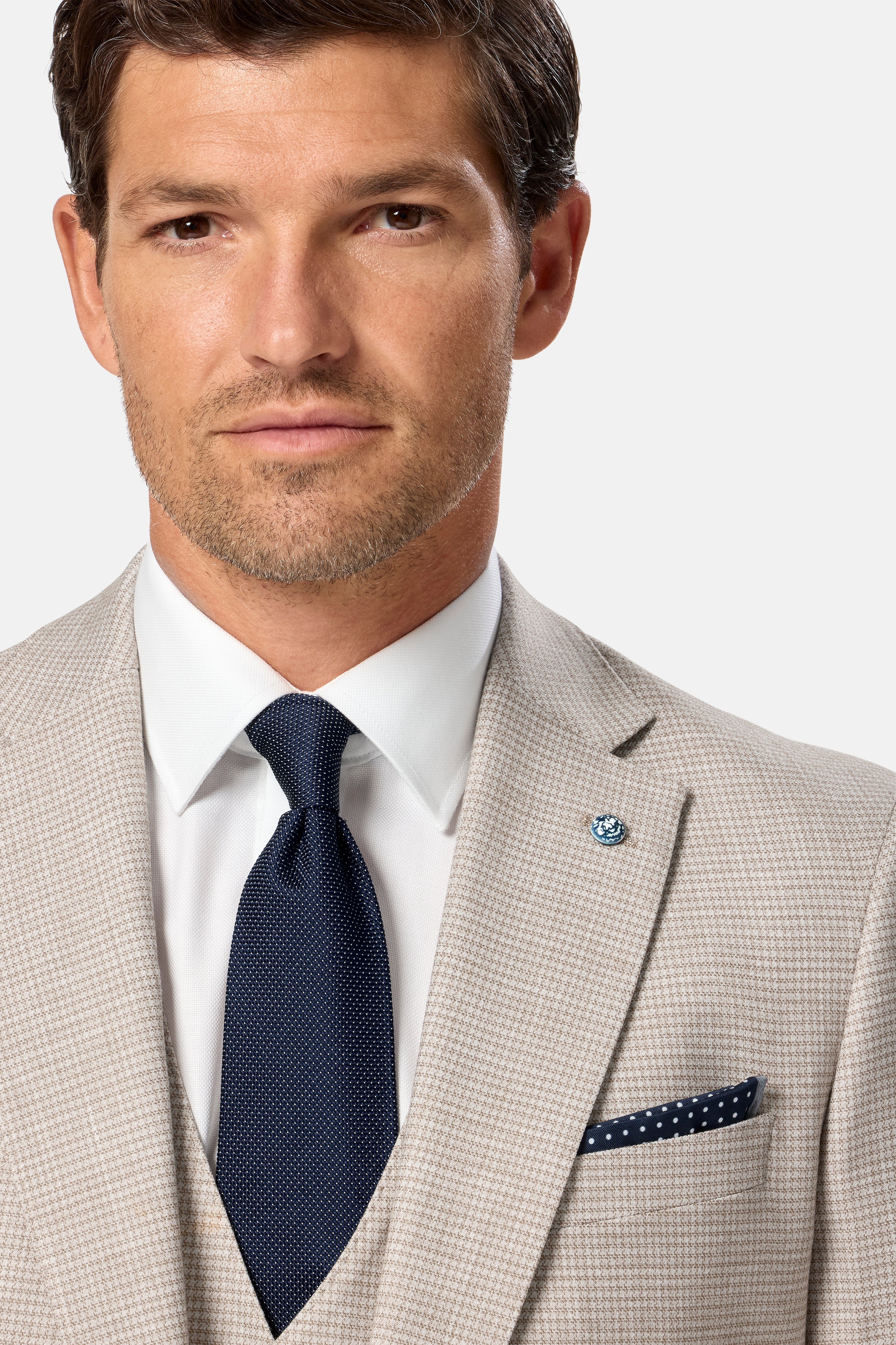 Man wearing a beige suit with a navy tie and pocket square on a light gray background