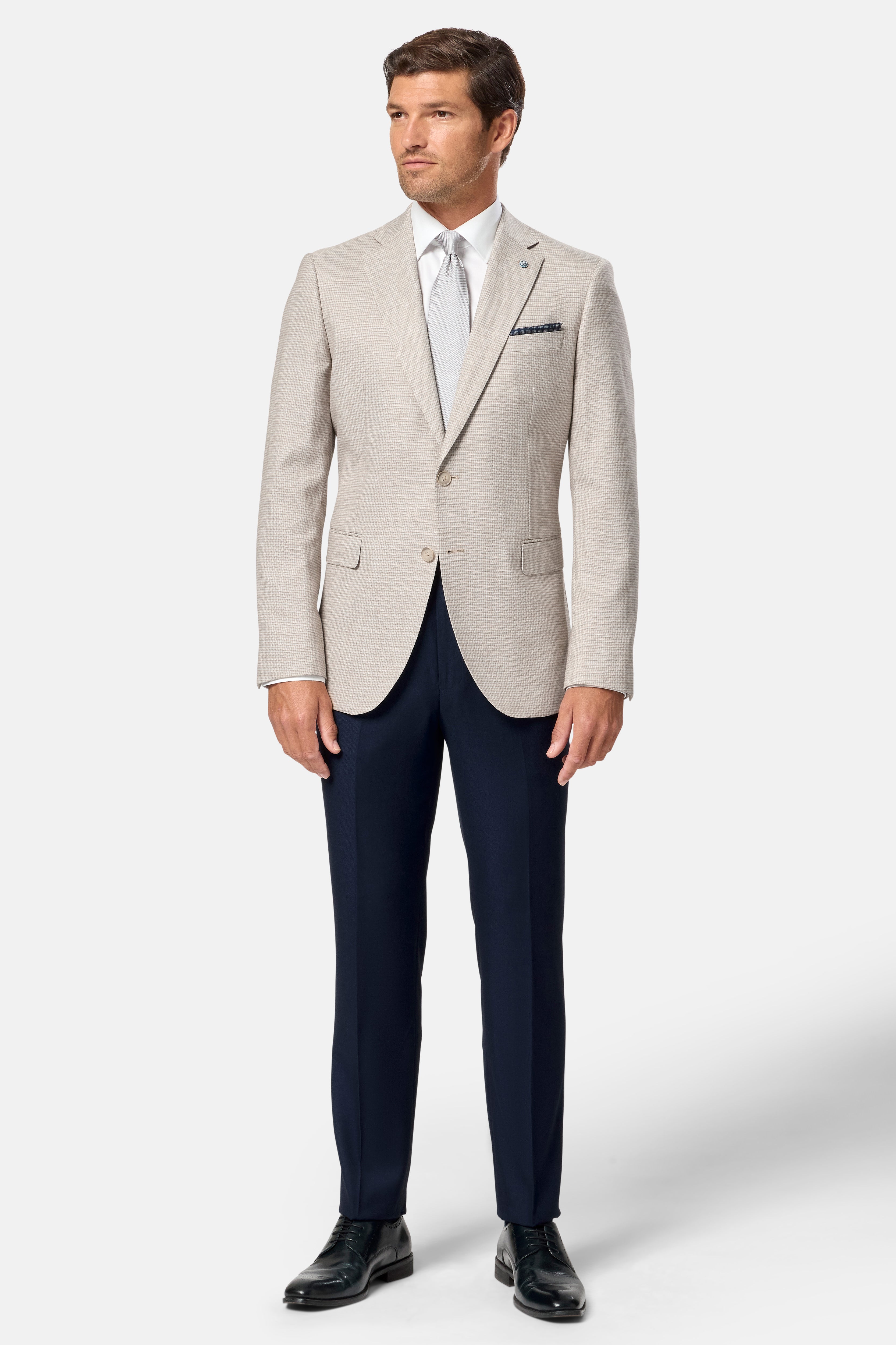 Man wearing a light stone blazer, navy pants, and black shoes on a white background