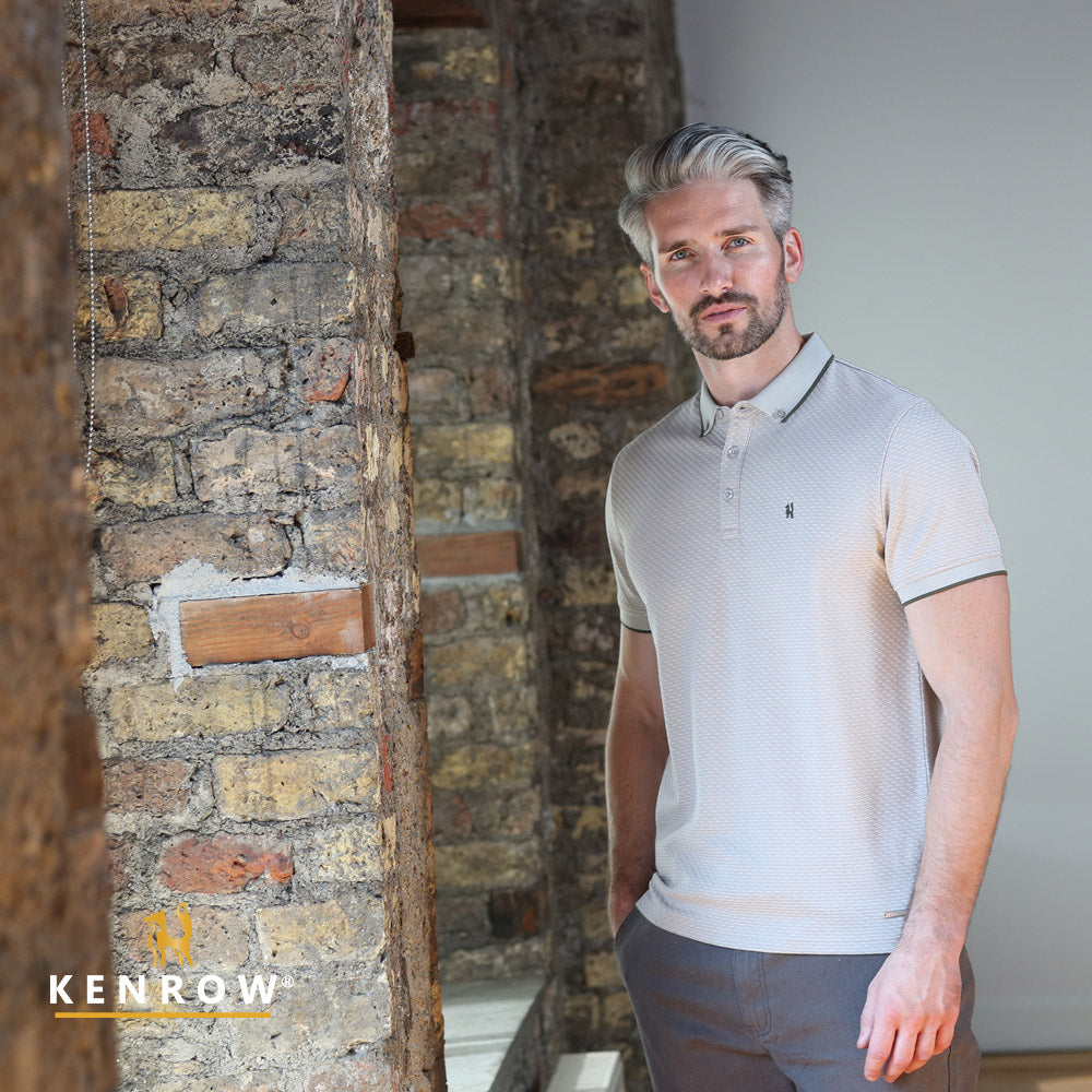 Man wearing a light gray polo shirt standing in a room with stone walls.