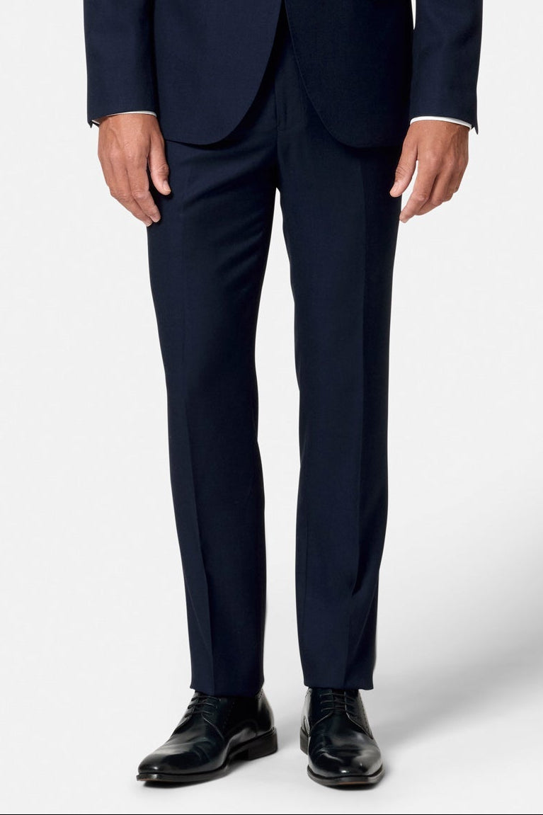 Man wearing a navy suit trousers with a white shirt and black shoes on a white background.