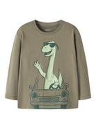 A long-sleeve crewneck t-shirt in a muted olive-brown color, featuring a cartoon graphic of a green dinosaur wearing sunglasses and driving a car.
