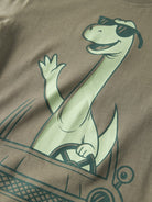 Close-up of a taupe-brown t-shirt featuring a cartoon graphic of a light green long-necked dinosaur wearing sunglasses and driving a car, with the dinosaur waving.