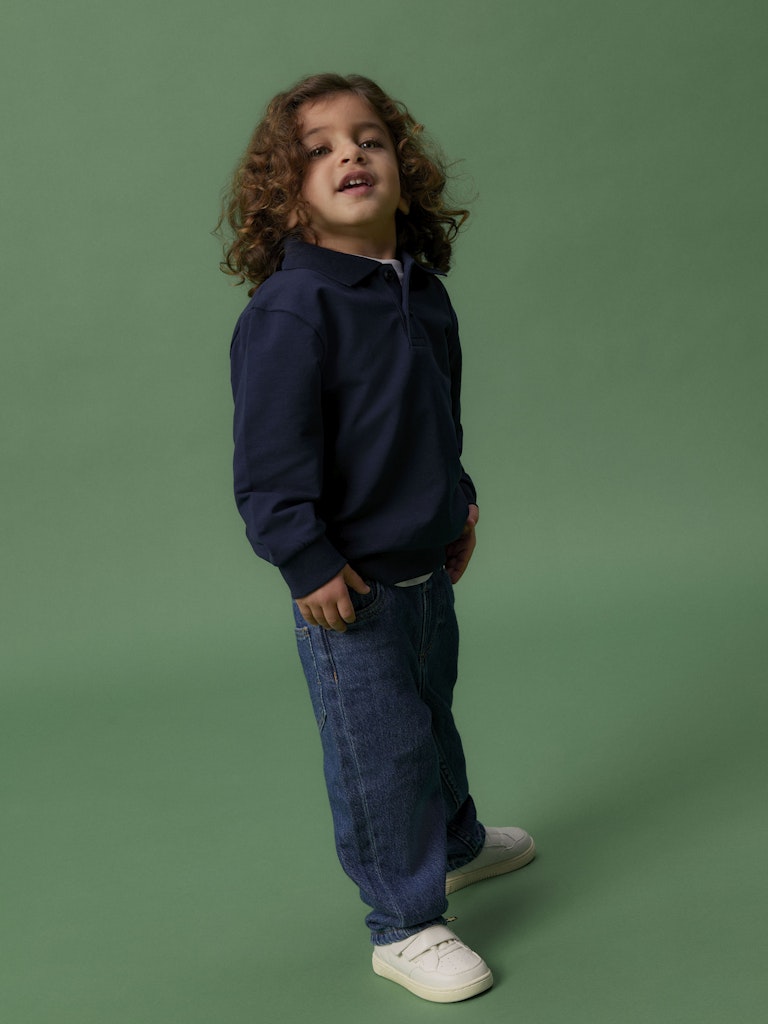 Child wearing a navy polo sweatshirt and blue jeans standing against a green background