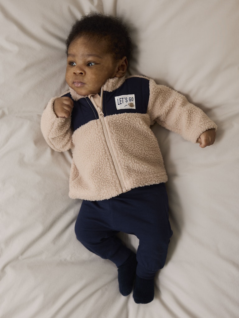 Baby wearing a beige fleece jacket with a visible brand logo on a white blanket