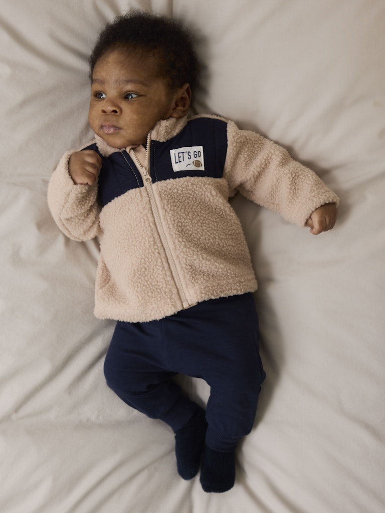 Baby wearing a beige and navy fleece outfit on a light-colored blanket