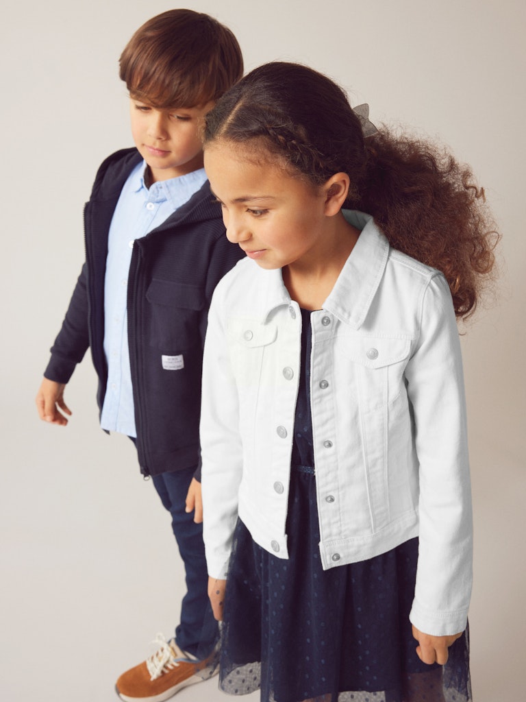 Two children standing side by side, one wearing a dark jacket and light shirt, the other in a white denim jacket and navy dress.