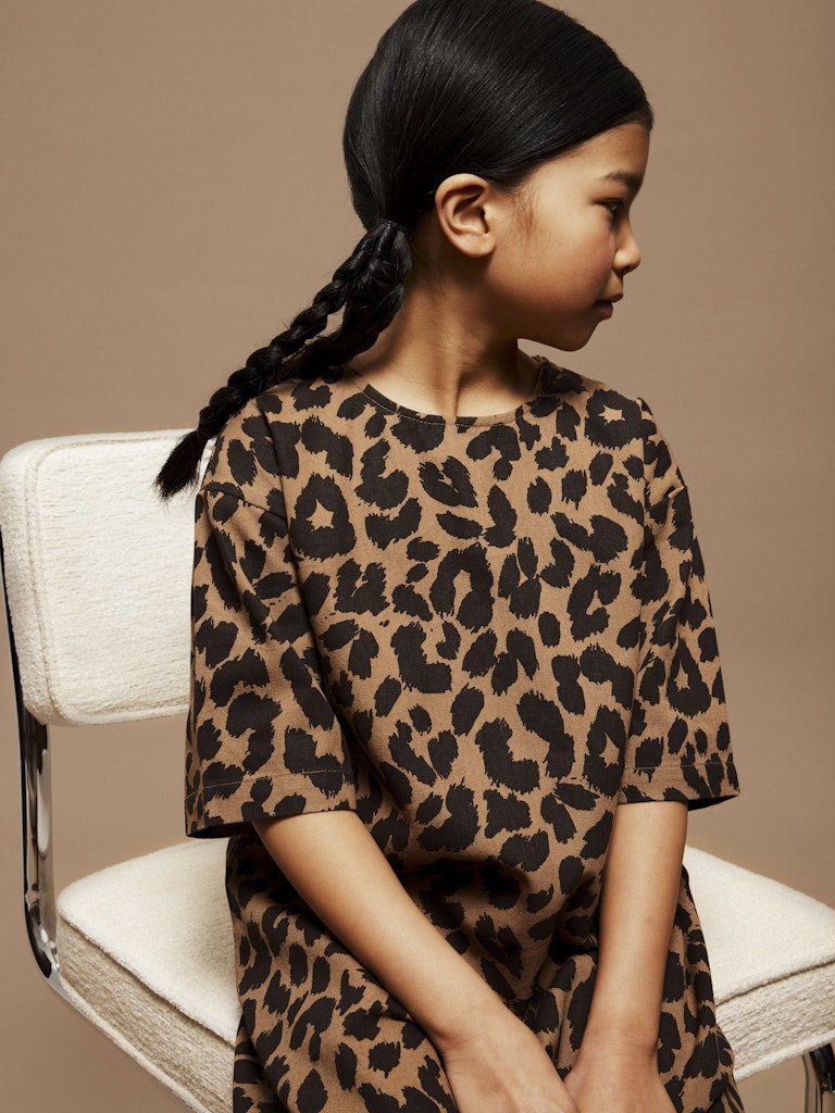 Young girl wearing a leopard print dress sitting on a chair against a beige background